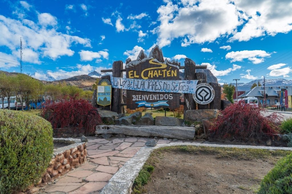 Cartel de madera de bienvenida a El Chaltén decorado con una bandera argentina bajo un cielo azul con nubes.