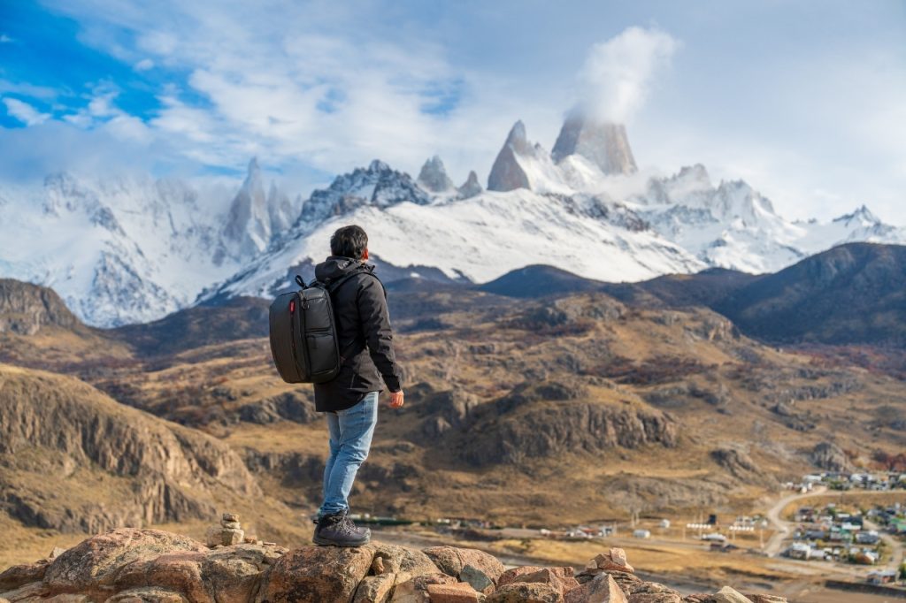 Un turista de espalda con mochila negra contemplando el imponente Cerro Fitz Roy nevado desde una cumbre rocosa sobre el valle.
