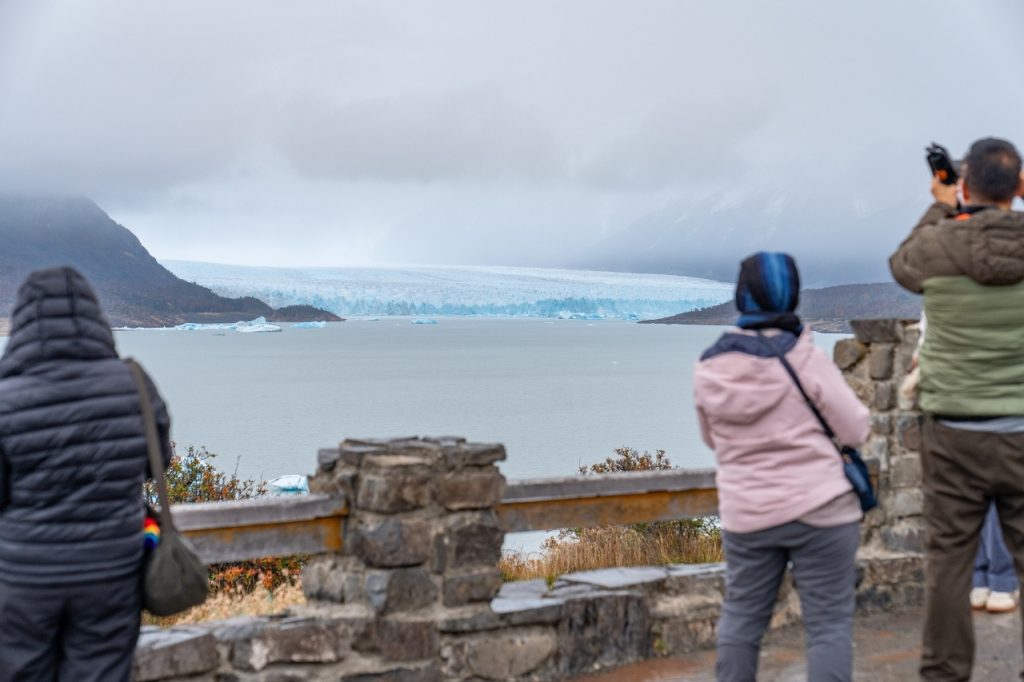 Turistas de espaldas junto a una baranda de piedra tradicional tomando fotografías del Glaciar Perito Moreno en un día nublado.