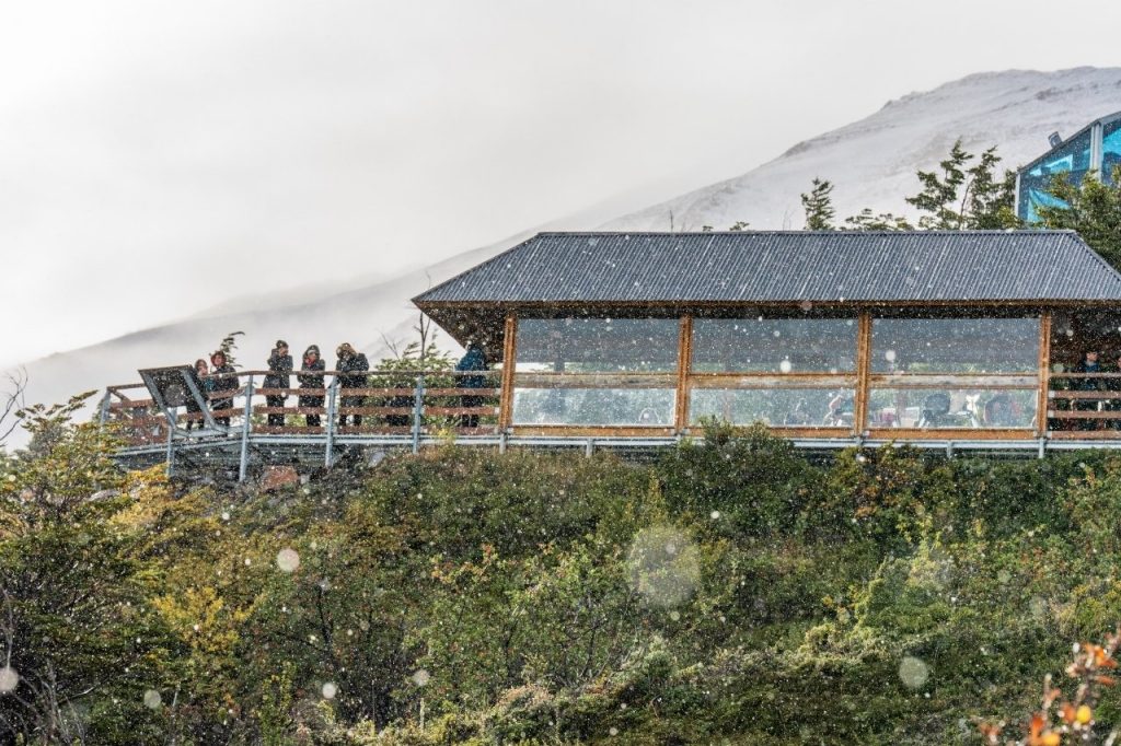 Turistas resguardados en un mirador de madera techado observando el paisaje del Parque Nacional Los Glaciares bajo una suave nevada.