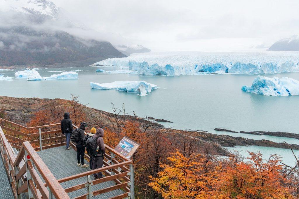Grupo de turistas con mochilas en un balcón de las pasarelas observando el Glaciar Perito Moreno y grandes témpanos de hielo flotando.