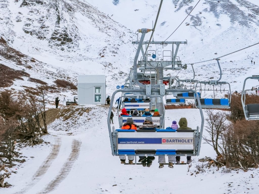 Pasajeros con ropa de invierno en primer plano de una silla de la aerosilla del Glaciar Martial, con montañas nevadas al fondo, Ushuaia.