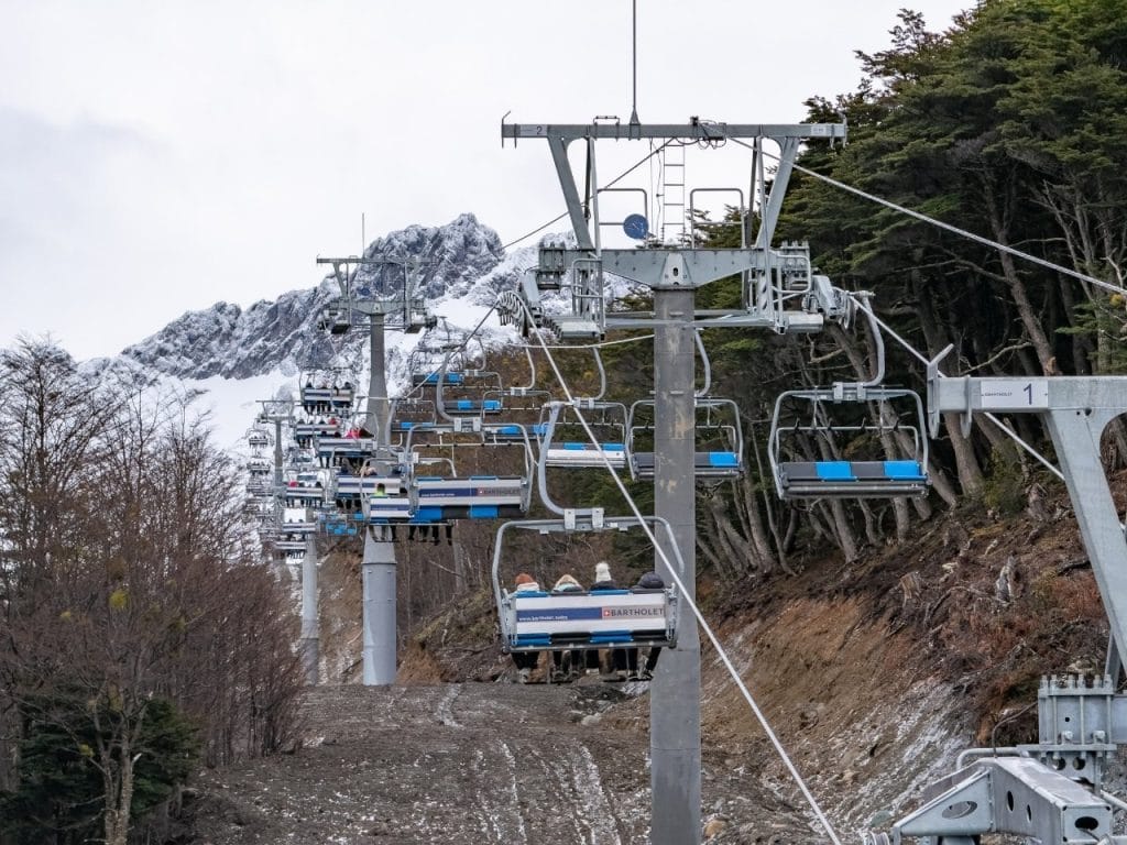 Primer plano de una silla de la aerosilla Barthollet con pasajeros, subiendo hacia el Glaciar Martial entre bosques y montañas nevadas en Ushuaia.