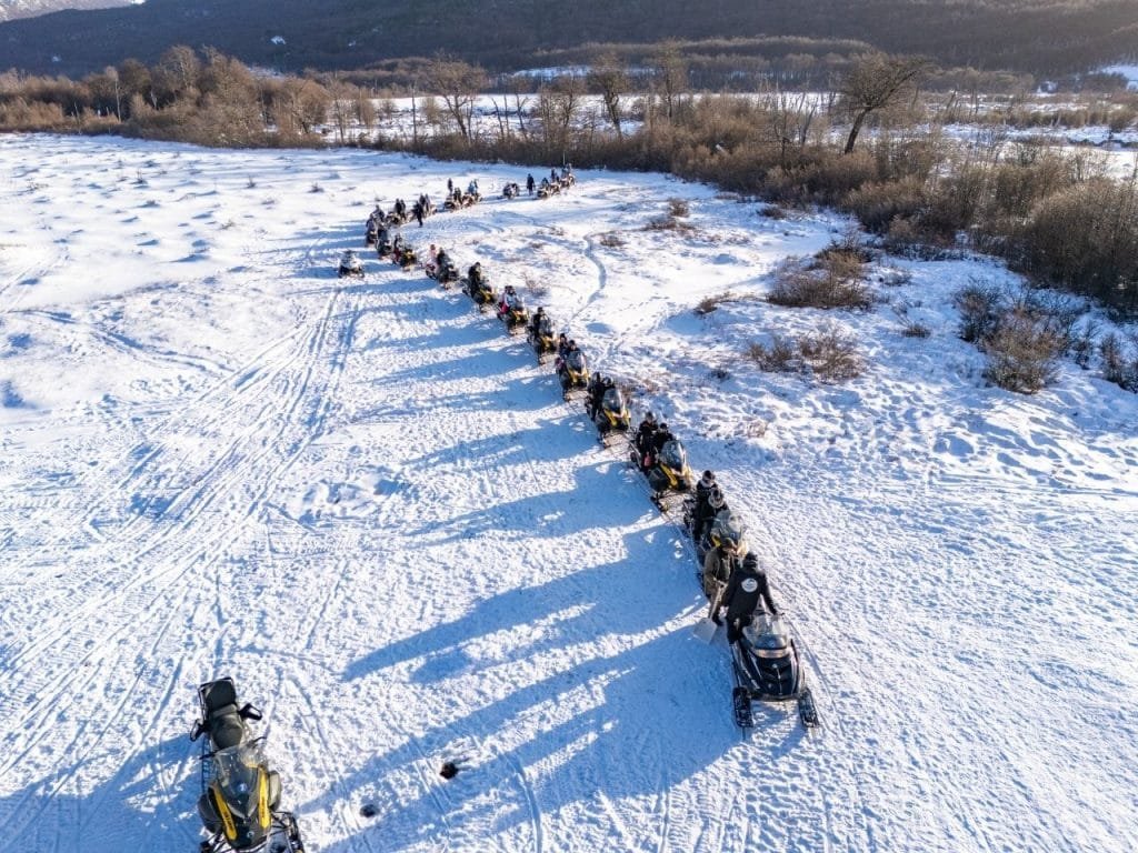 Fotografía de dron que muestra varias motos de nieve circulando por caminos trazados en la nieve profunda del Park Austral, con montañas de fondo.