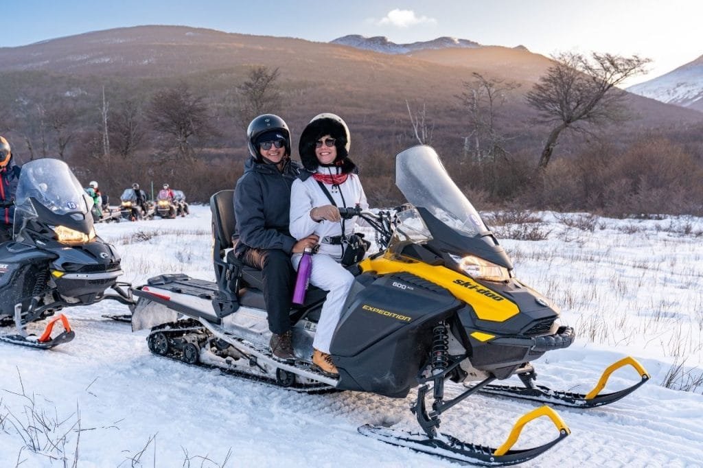 Una joven sonriente se desliza en un trineo de plástico rosa por una colina nevada en el Park Austral, con otras personas y una red de seguridad de fondo.